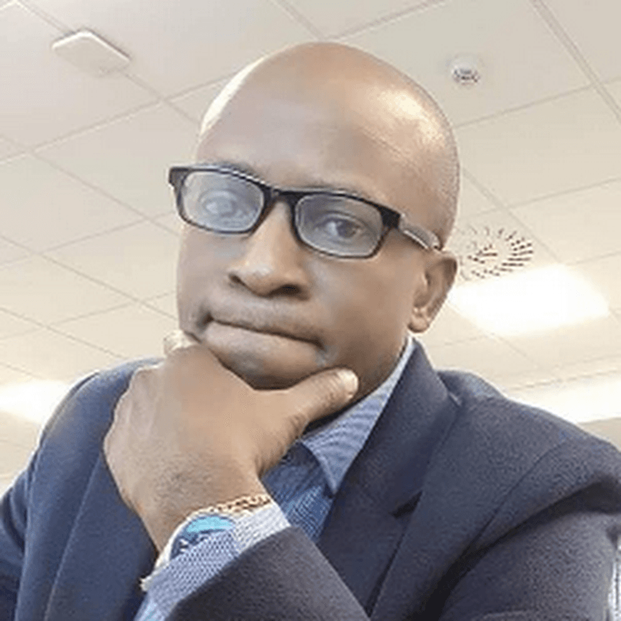Bald Black man in glasses and a blazer, thoughtfully resting his chin on his hand.